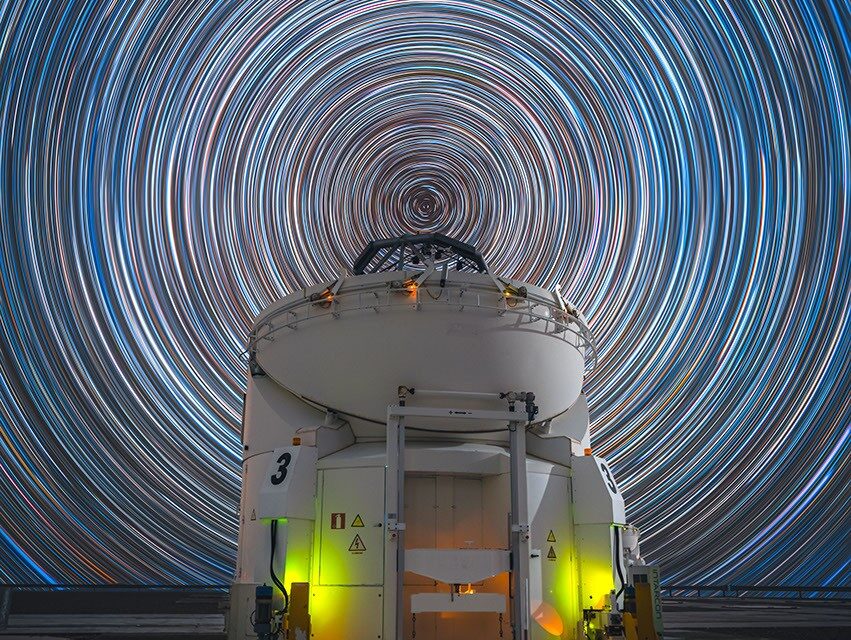 Rastros de estrelas concêntricos ao redor do polo celeste sul sobre o Telescópio Auxiliar AT 3 no Observatório de Paranal, Chile.